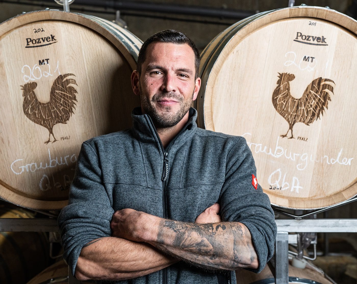 Ein Mann mit Tätowierungen auf den Unterarmen steht mit verschränkten Armen vor großen hölzernen Weinfässern, die jeweils mit einem Hahn und handgeschriebenen Etiketten versehen sind. Er trägt eine graue Jacke mit Reißverschluss und lächelt selbstbewusst.