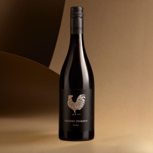 Eine Rotweinflasche mit schwarzem Etikett, auf dem ein Hahn und der Text "CABERNET SAUVIGNON" abgebildet sind, steht aufrecht auf einer beigen Fläche vor einem sanft geschwungenen, beige-braunen Hintergrund.