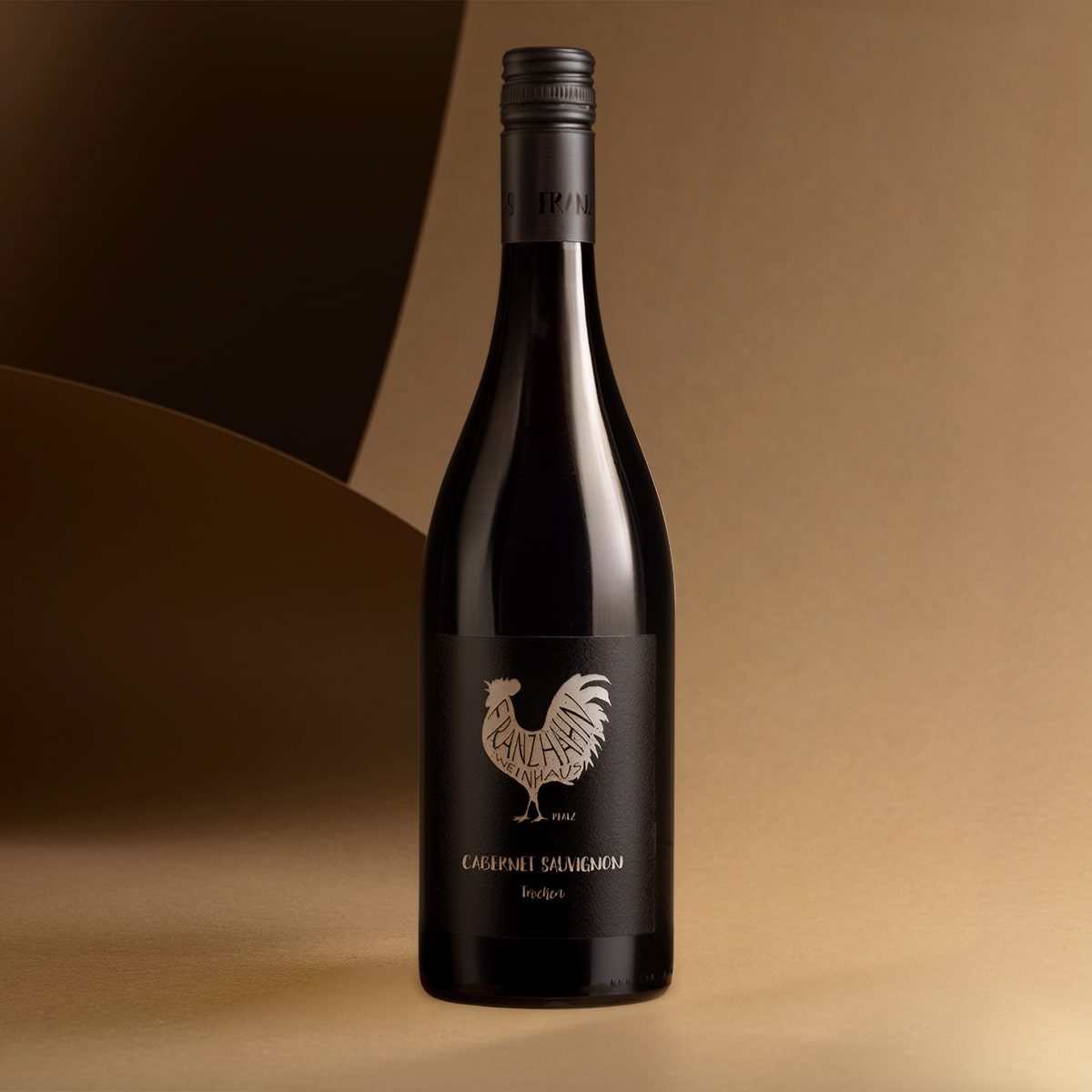 Eine Rotweinflasche mit schwarzem Etikett, auf dem ein Hahn und der Text "CABERNET SAUVIGNON" abgebildet sind, steht aufrecht auf einer beigen Fläche vor einem sanft geschwungenen, beige-braunen Hintergrund.