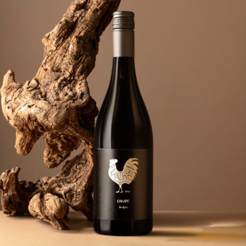Eine dunkle Weinflasche mit silbernem Verschluss und weißem Hahn-Logo auf dem Etikett steht neben einem großen Stück gedrehten Treibholzes auf einem beigen Hintergrund.
