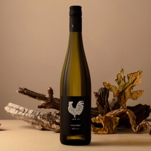 Eine Weißweinflasche mit einem schwarzen Etikett mit einem Hahn-Logo auf einer beigen Fläche mit getrocknetem Holz und Blättern als Dekoration im Hintergrund. Die Umgebung ist warm und natürlich beleuchtet.