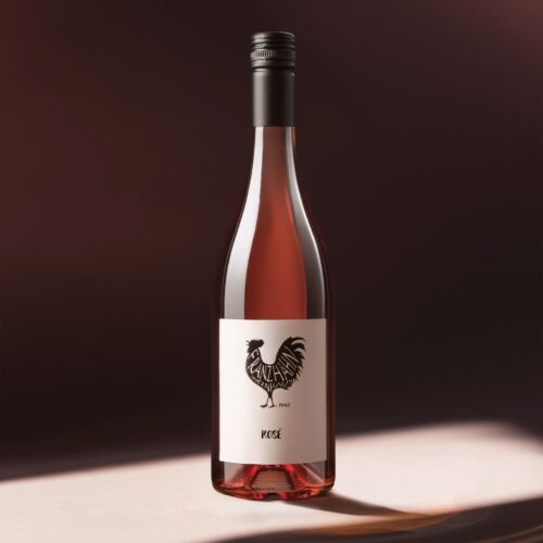 Eine Flasche Roséwein mit einem dunklen Schraubverschluss steht auf einer Fläche. Das weiße Etikett zeigt die schwarze Illustration eines Hahns und die Worte "NATURAL WINE" und "ROSÉ". Der Hintergrund ist sanft in warmen Tönen beleuchtet.