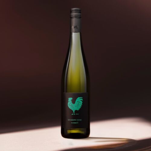 Eine große grüne Weinflasche mit einem schwarzen Etikett mit türkisfarbener Hahngrafik und weißem Text mit der Aufschrift "Sauvignon Blanc Rudeine" steht auf einer Fläche mit dramatischer Beleuchtung und einem dunklen, verschwommenen Hintergrund.