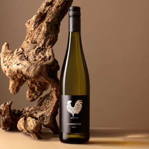 Eine Weißweinflasche mit einem schwarzen Etikett, auf dem ein weißer Hahn abgebildet ist, steht aufrecht auf einer beigen Fläche, neben einem großen, strukturierten Stück Treibholz vor einem neutralen Hintergrund.