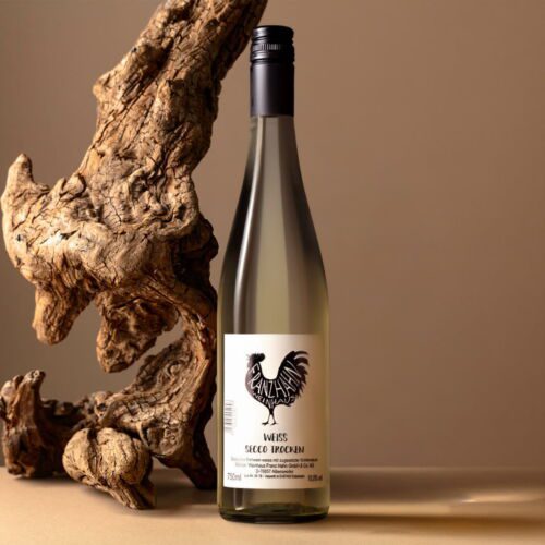 Eine Flasche Weißwein Weiss Secco steht auf einer beigen Fläche neben einem großen, strukturierten Stück Treibholz vor einem neutralen Hintergrund.