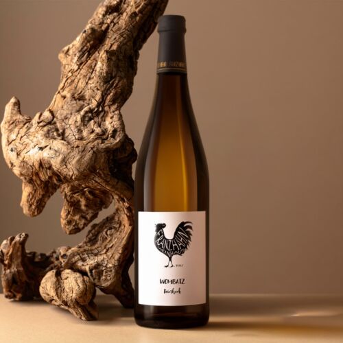 Eine Flasche Weißwein mit einem Hahn-Logo auf dem Etikett steht auf einer flachen Oberfläche neben einem großen, strukturierten Stück Treibholz vor einem beigen Hintergrund.