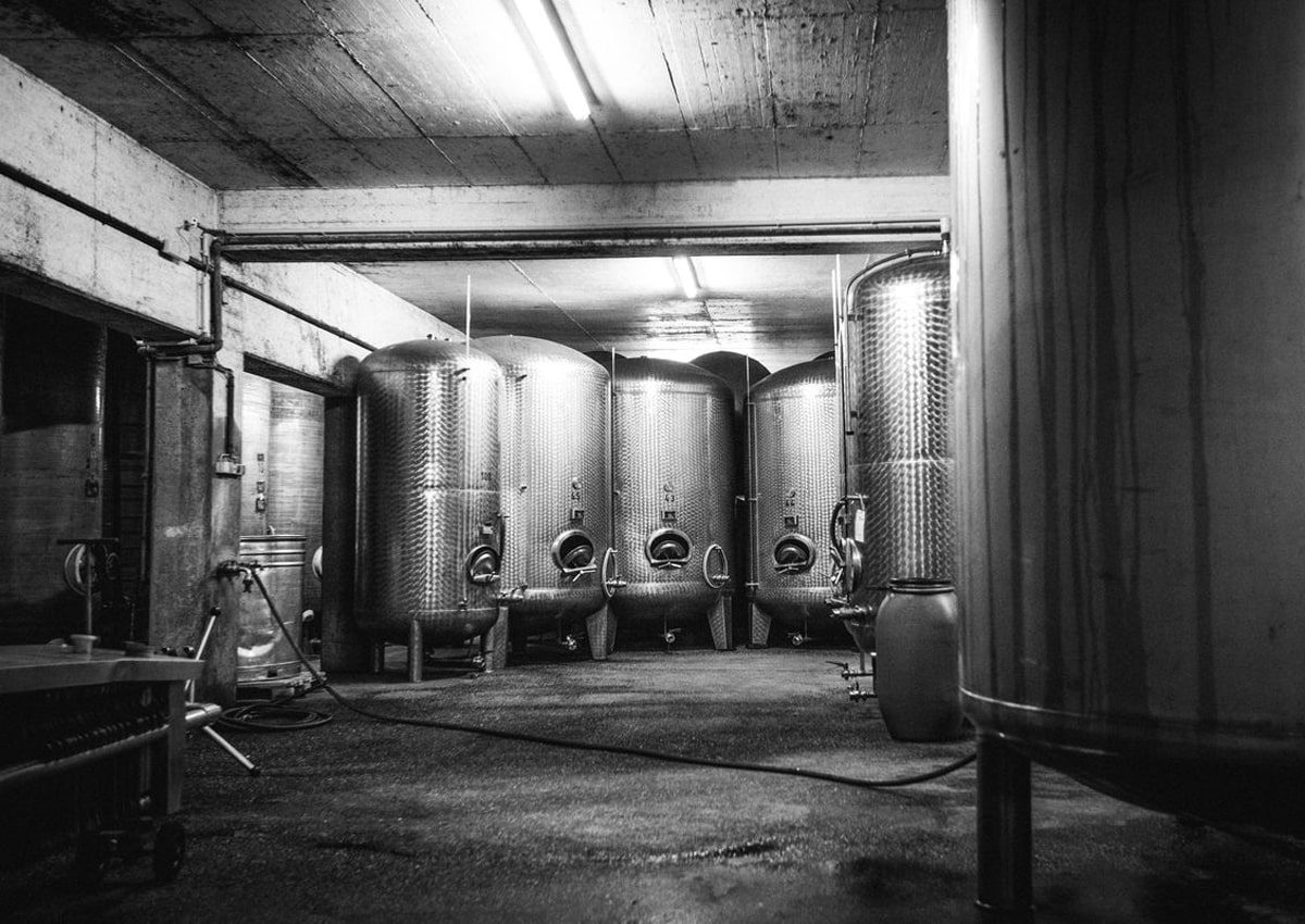 Schwarz-Weiß-Foto eines Brauerei- oder Brennereiraums mit mehreren großen Gärtanks aus Metall, die an der Rückwand aufgereiht sind, Schläuchen auf dem Boden und einem Fass im Vordergund. Von der Decke hängen Leuchtstoffröhren.