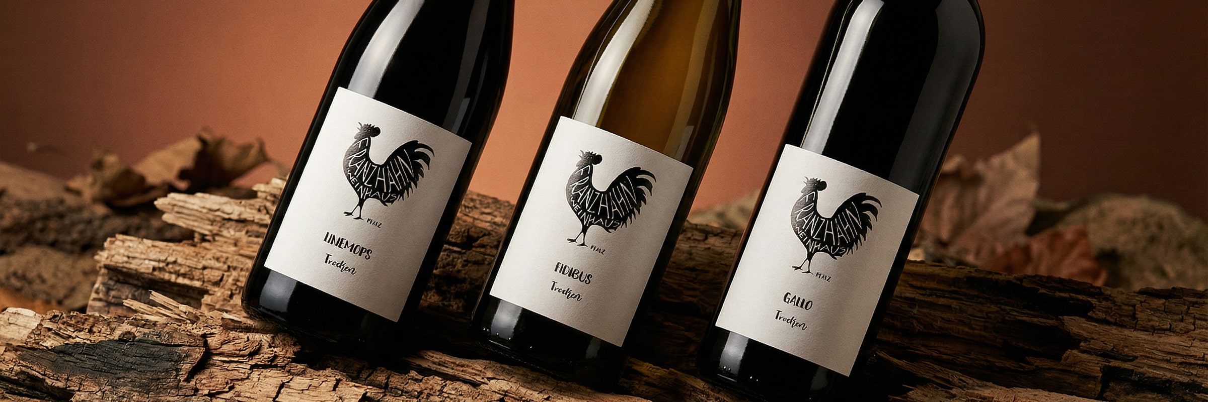 Drei Weinflaschen mit schwarz-weißen Etiketten, auf denen ein stilisierter Hahn abgebildet ist, stehen auf einer rustikalen Holzfläche mit trockenen Blättern im Hintergrund.