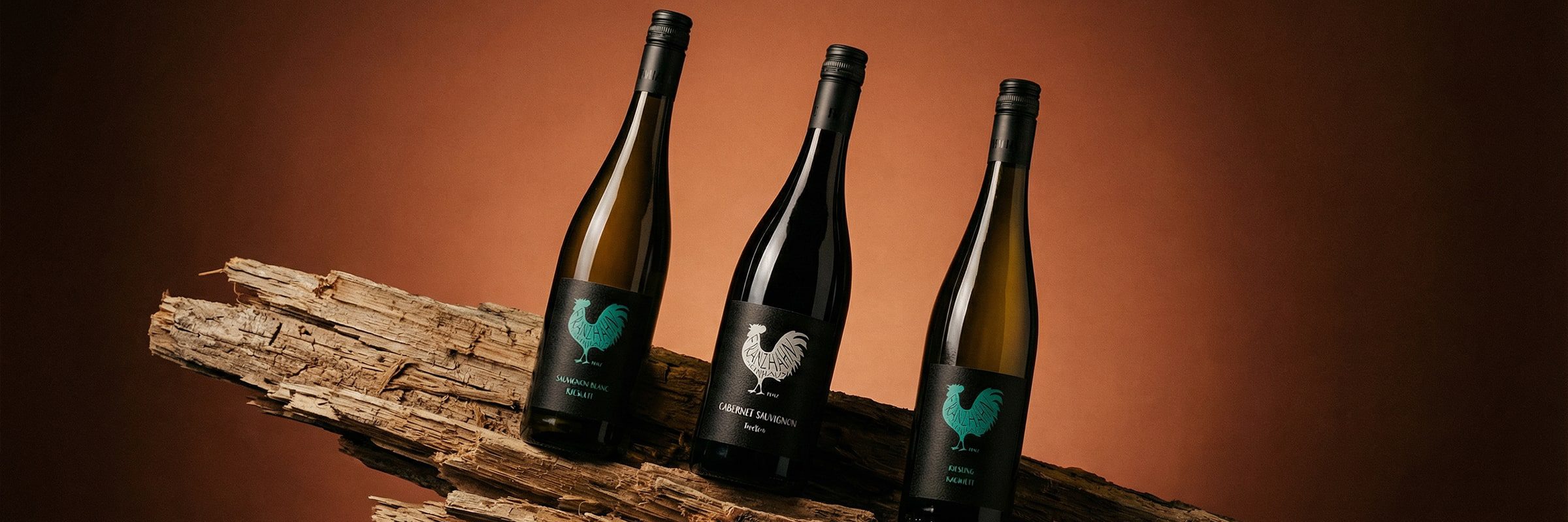 Drei Weinflaschen mit schwarzen Etiketten, auf denen ein Hahn abgebildet ist, ruhen auf einem Stück rauen Holzes vor einem Hintergrund mit warmem braunen Farbverlauf.
