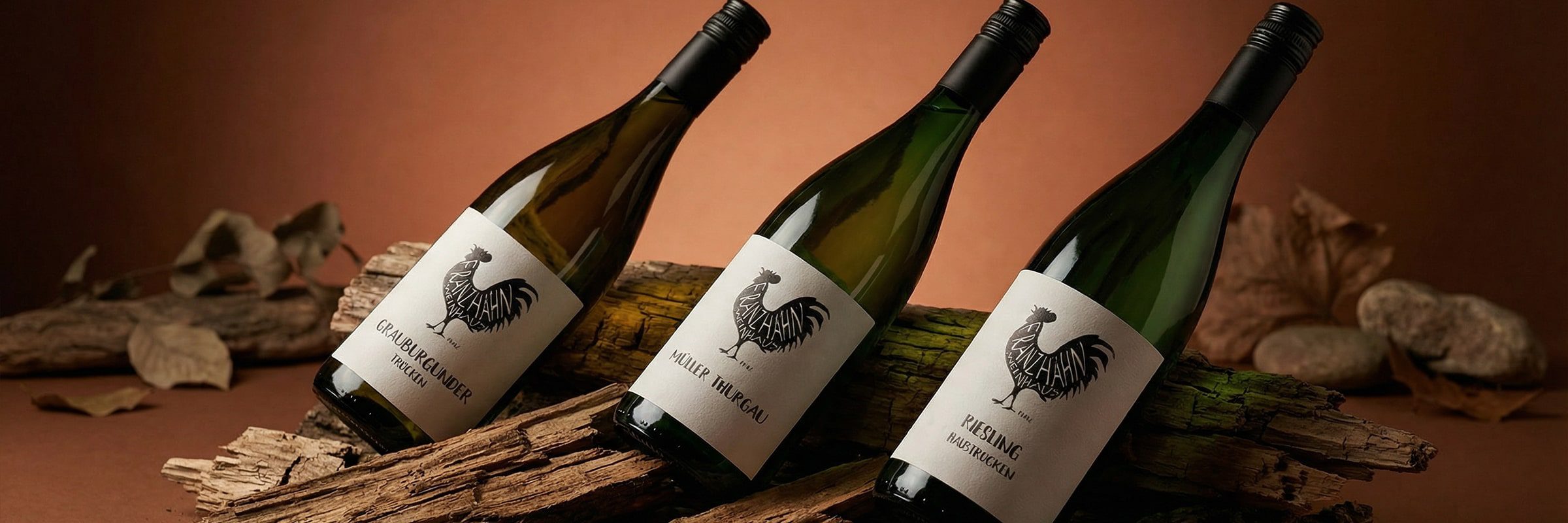 Drei Weinflaschen mit dem Logo eines Hahns auf dem Etikett liegen auf einem bemoosten Baumstamm, umgeben von getrockneten Blättern und Steinen, vor einem braunen Hintergrund.