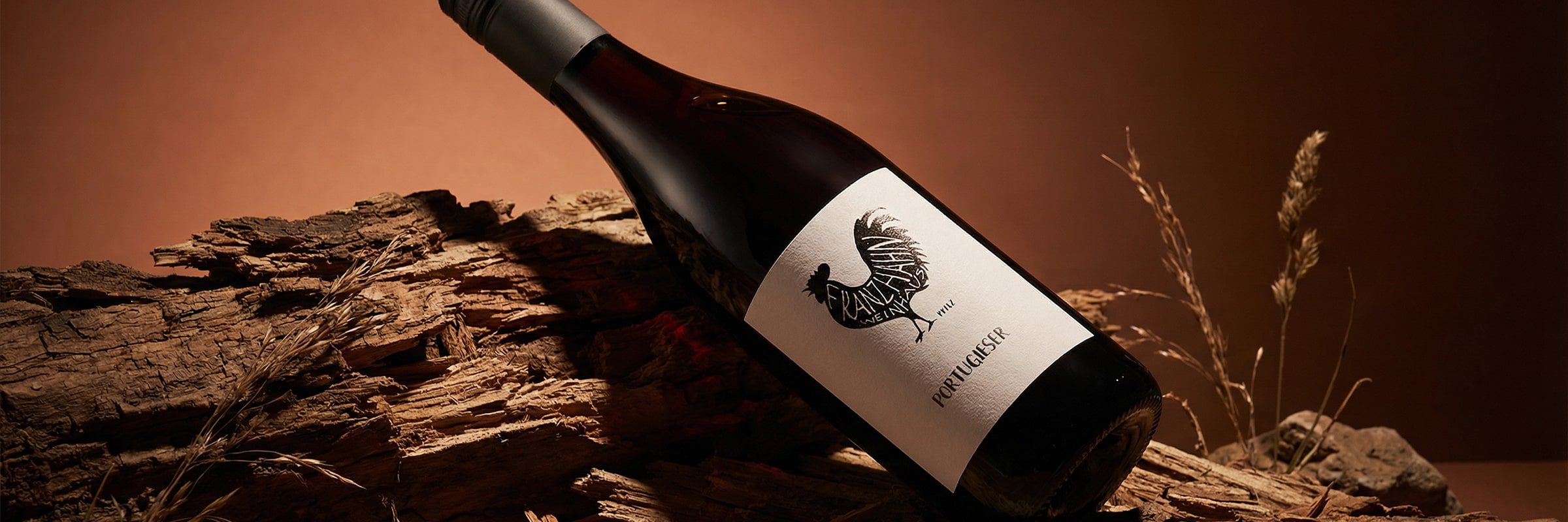 Eine Rotweinflasche mit der Aufschrift "Portugieser" liegt auf einem Stück rustikalen Holzes vor einem warmen braunen Hintergrund mit getrockneten Gräsern, der eine erdige, elegante Atmosphäre schafft.
