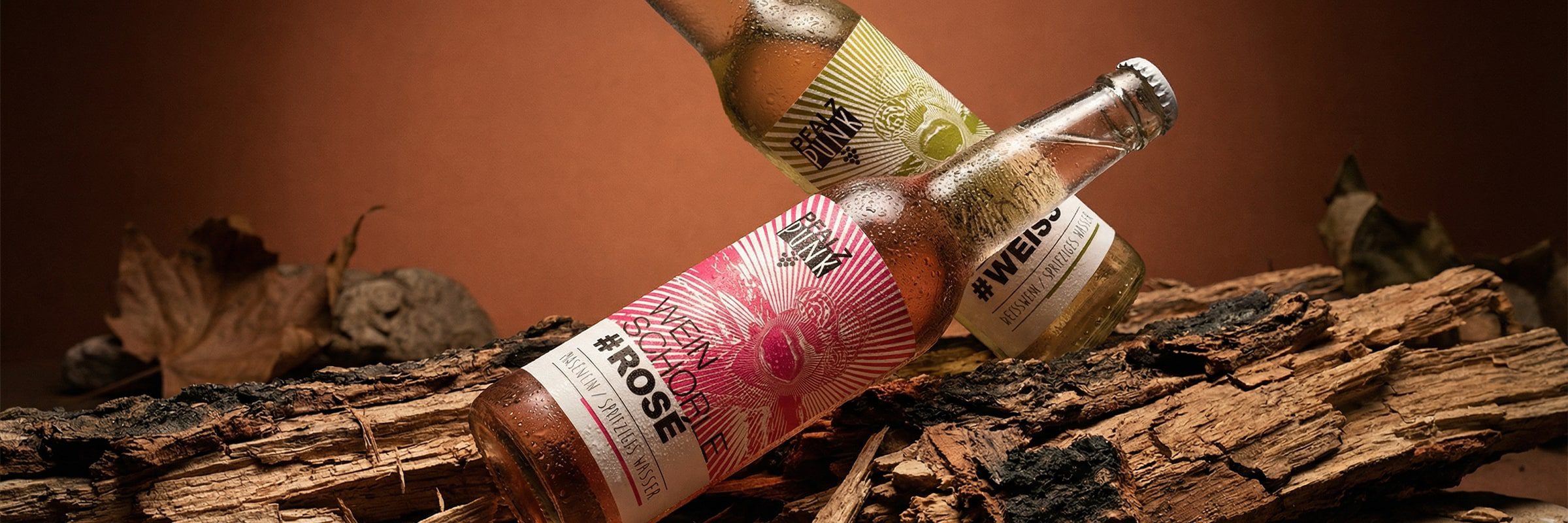 Zwei Glasflaschen mit kohlensäurehaltigen Getränken, die mit "#ROSE" und "#WEISS" beschriftet sind, liegen auf einem Stück rauen Holzes, wobei getrocknete Blätter und ein warmer brauner Hintergrund eine rustikale Herbstatmosphäre schaffen.