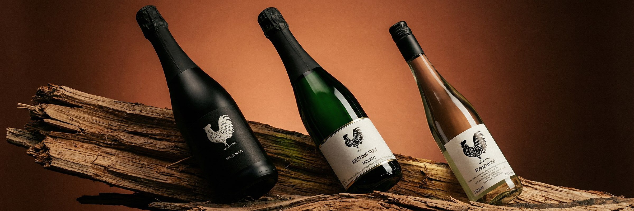 Drei Weinflaschen, jede mit einem Hahn-Logo, sind auf einem Stück verwitterten Holzes vor einem Hintergrund mit braunem Farbverlauf ausgestellt. Die Flaschen sind schräg angeordnet, mit verschiedenfarbigen Etiketten und Verschlüssen.