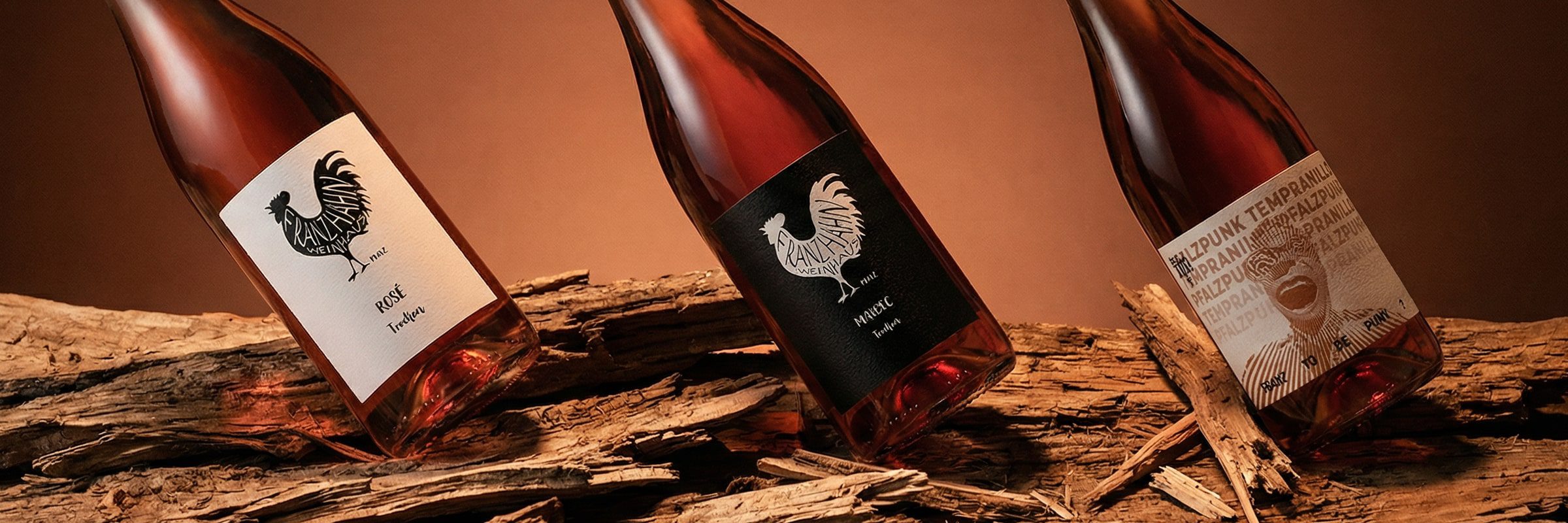 Drei Flaschen Roséwein mit Hahn-Etiketten sind schräg auf rustikalen Holzteilen vor einem warmen braunen Hintergrund ausgestellt.