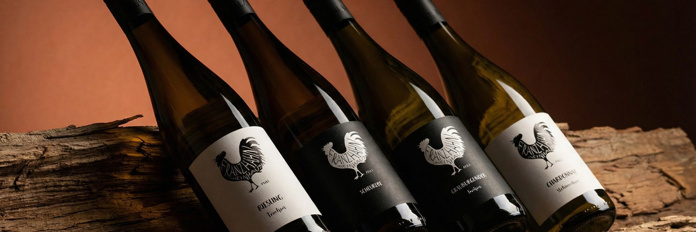 Vier Weinflaschen mit Hahnlogos und verschiedenen Etiketten stehen auf einem Stück rustikalem Holz vor einem warmen braunen Hintergrund.