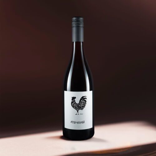 Eine Rotweinflasche von Assemblage mit einem schwarzen Schraubverschluss steht auf einer hellen Fläche. Auf dem weißen Etikett ist ein schwarzer Hahn abgebildet, darunter steht der Name "Assemblage" vor einem dunklen Hintergrund mit Farbverlauf.