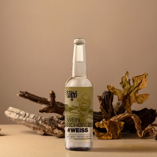 Eine Glasflasche von Schorle Weiss steht auf einer beigen Fläche, auf der getrocknetes Holz und große braune Blätter als natürliche Kulisse vor einem hellbraunen Hintergrund angeordnet sind.