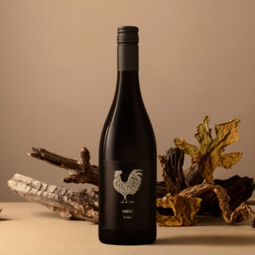 Eine Flasche Shiraz mit einem schwarzen Etikett, auf dem ein Hahn abgebildet ist, steht auf einer beigen Fläche, umgeben von getrockneten Blättern und Holzstücken, vor einem neutralen Hintergrund.
