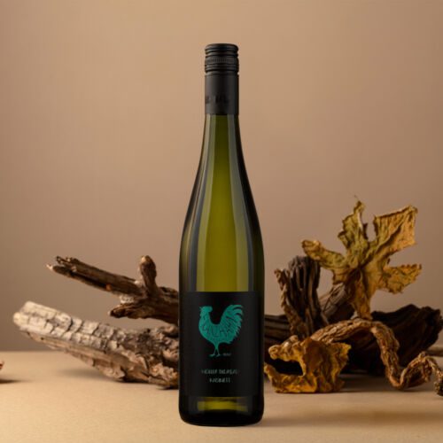 Eine grüne Müller-Thurgau Kabi Weinflasche mit schwarzem Etikett, auf dem ein türkisfarbener Hahn abgebildet ist, steht aufrecht auf einer beigen Fläche, umgeben von trockenem Treibholz und Herbstlaub im Hintergrund.